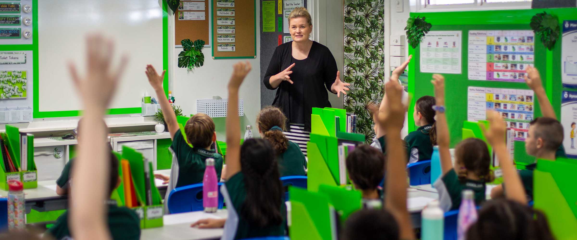 An educator standing at the front of a classroom, several children have their hands raised to ask a question.
