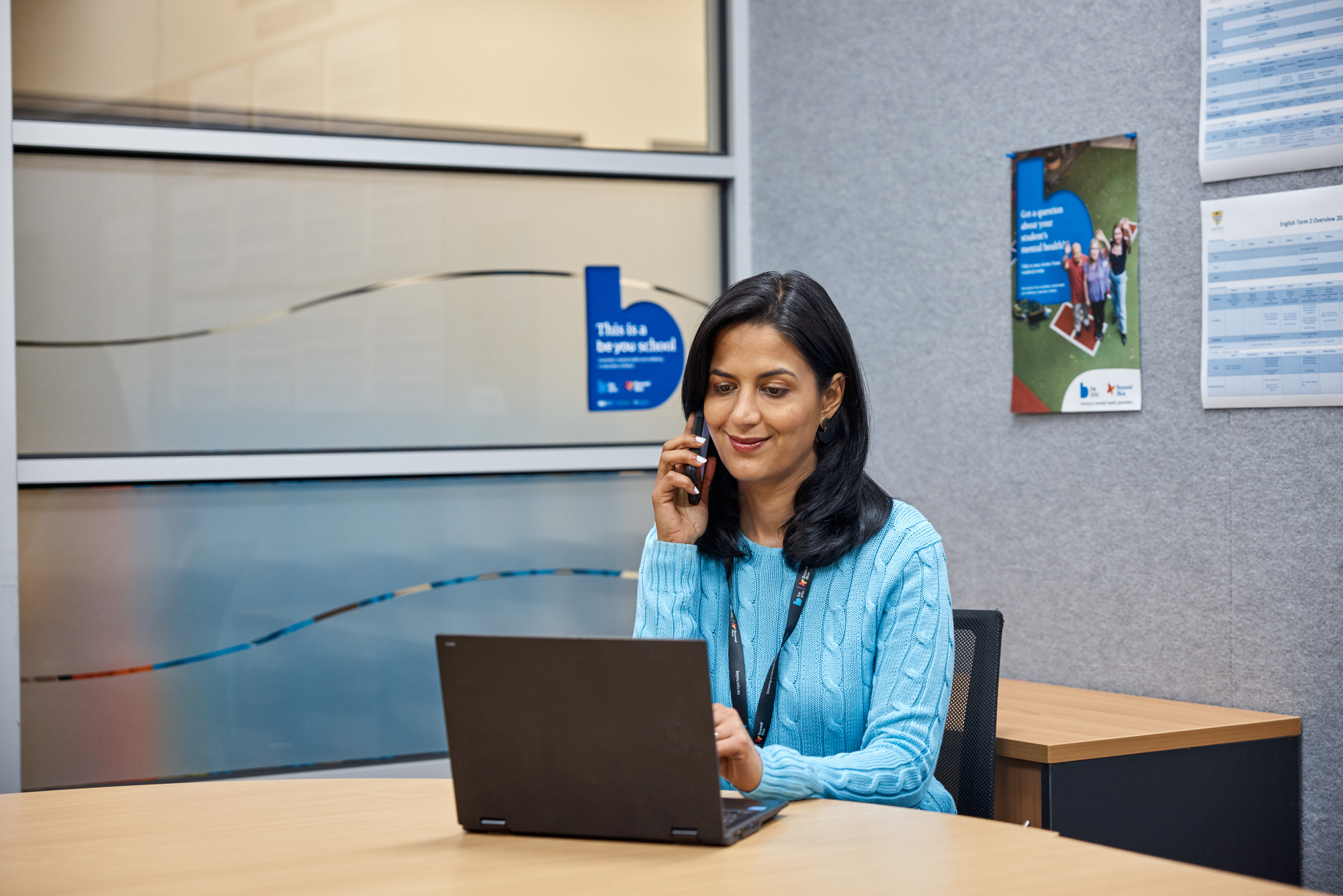 The image shows a female Be You Consultant sitting at a desk in an office setting, using a laptop computer and talking on a cell phone. She is wearing a blue sweater and appears to be engaged in a conversation. The office environment includes a large window, desk, and various informational posters on the wall. This image could be used to describe a professional working in an learning community.