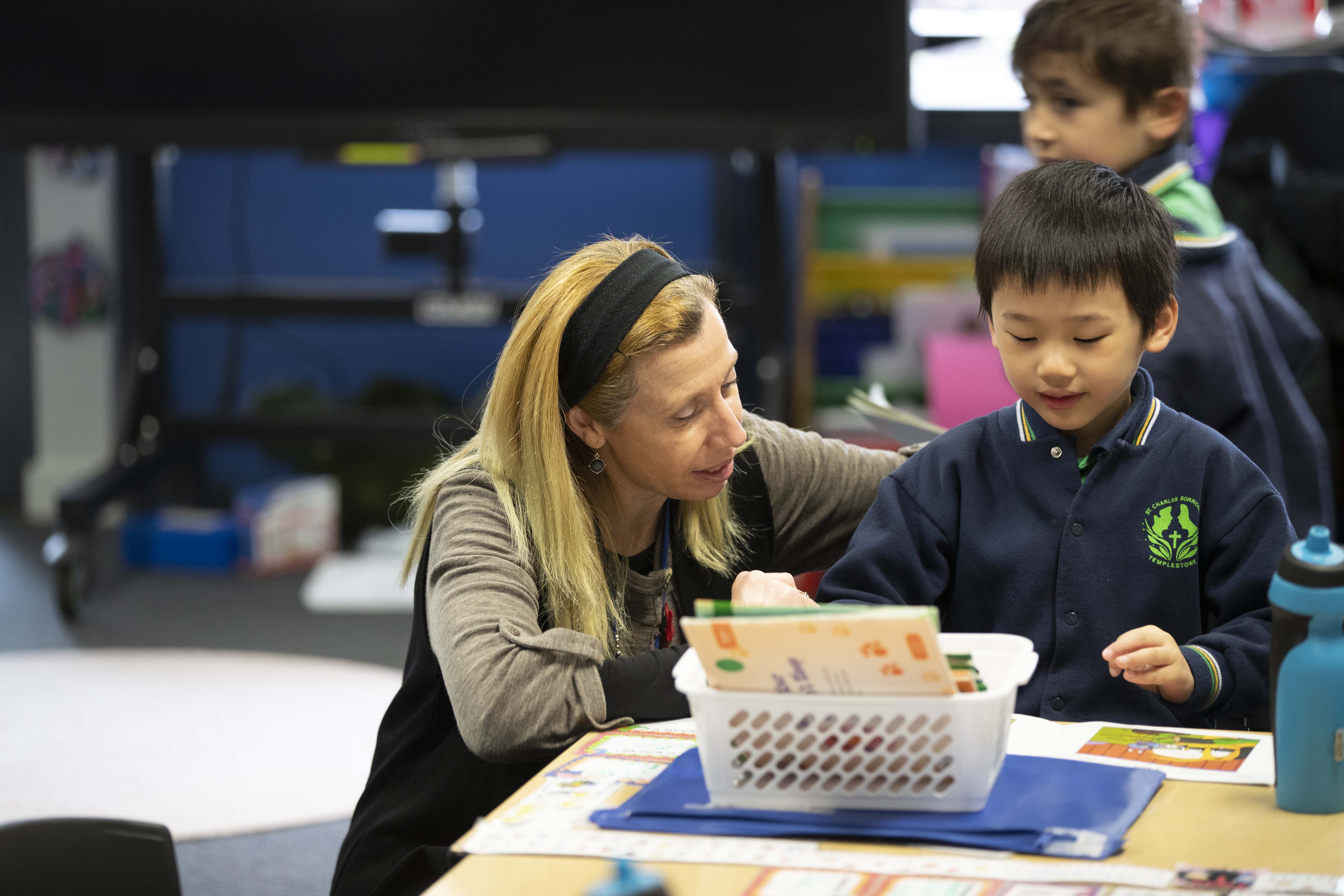  teacher kneeling down at a desk next to a primary school aged student.
